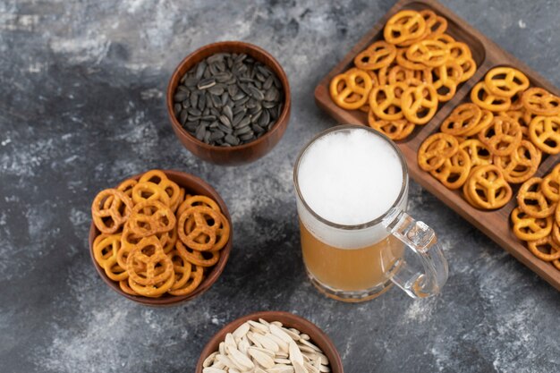 Bowls of salted pretzels, black sunflower seeds and mug of beer on marble surface.