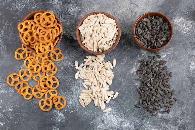 Bowls of salted pretzels, white and black sunflower seeds on marble surface