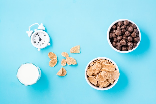 Bowls with cereal flakes and chocolate balls, a glass of milk and an alarm clock on a blue table. Scheduled breakfast, choice of dishes. Top view