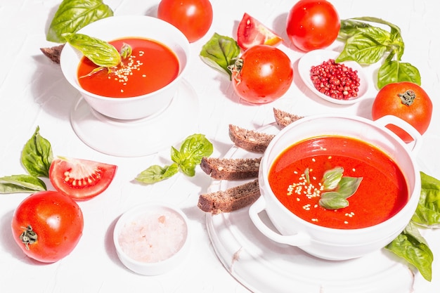 Bowls with homemade tomato soup. Ripe vegetables, fresh basil leaves, breadsticks, aromatic spices. Trendy hard light, dark shadow. White putty background, flat lay