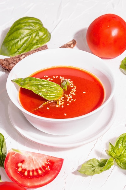 Bowls with homemade tomato soup. Ripe vegetables, fresh basil leaves, breadsticks, aromatic spices. Trendy hard light, dark shadow. White putty background, flat lay