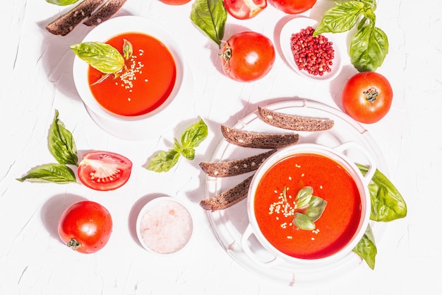 Bowls with homemade tomato soup. Ripe vegetables, fresh basil leaves, breadsticks, aromatic spices. Trendy hard light, dark shadow. White putty background, top view