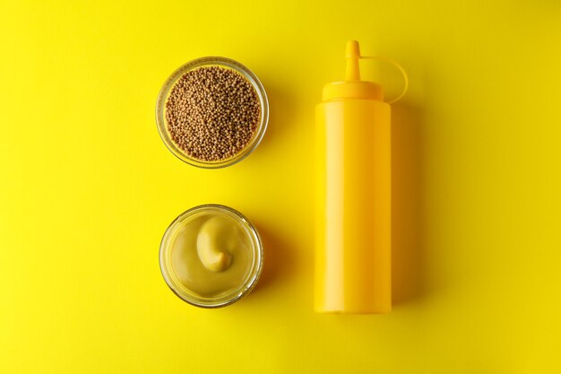 Bowls with mustard and sauce bottle on yellow background