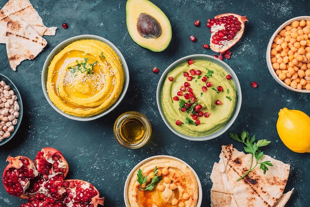 bowls with yellow hummus and green hummus tahini olive oil sesame seeds pita raw chickpeas avocado pomegranate on dark background middle eastern jewish arabic cuisine top view