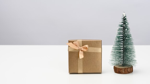 상자는 갈색 종이로 싸여 있고 흰색 테이블에 장식용 크리스마스 트리가 있습니다. 축제 배경, 복사 공간