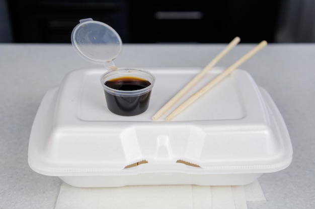 A box on a light table at home in the kitchen Fast delivery sushi in a white container Soy sauce jar and sushi sticks