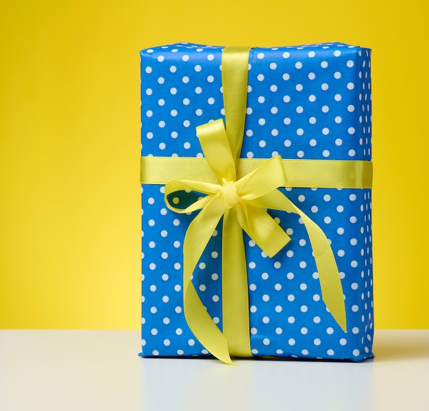 Box packed in festive blue paper and tied with silk yellow ribbon on a yellow background, birthday gift, surprise