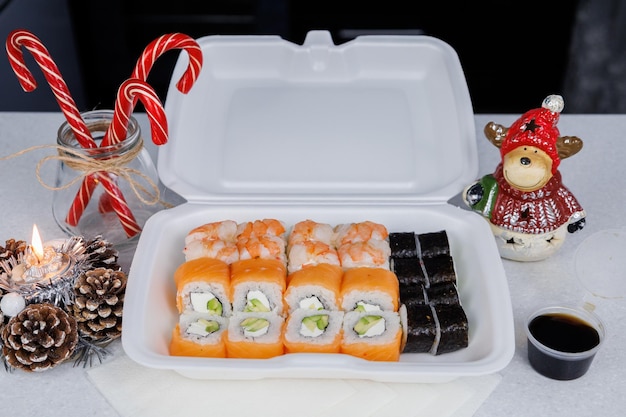 부엌에 있는 테이블에 있는 필라델피아 롤 한 상자 흰색 용기에 담긴 빠른 배달 스시 스시를 위한 간장 한 병 축제 장식 크리스마스 컨셉