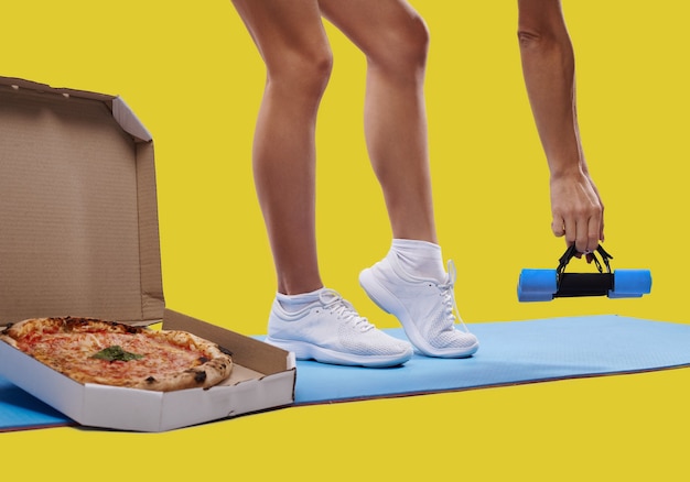 おいしい新鮮なピザの箱、ヨガマットの上に認識できないフィットの女性の脚、そして隔離されたダンベルを持っている手。体重を減らし、太った概念を取得します。フィットネスとダイエットのコンセプト。