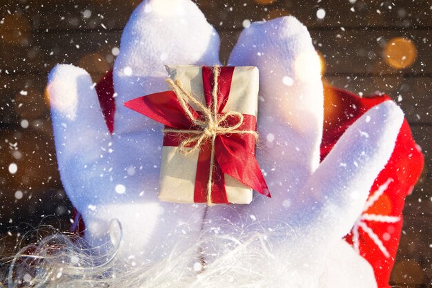 흰색 장갑을 낀 산타클로스의 손에 크리스마스 선물이 든 상자. 빨간 양복, 수염, 화환 조명이 흐릿합니다. 새해, 준비, 기적에 대한 기대, 꿈이 이루어집니다. 확대