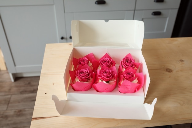 Box with creamy cupcakes decorated with rose buds on the table, desserts delivery concept.