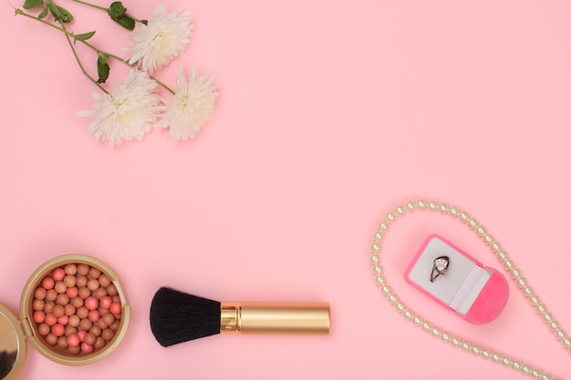 Box with ring, beads, powder, brush and flowers on a pink background. Women cosmetics and accessories. Top view with copy space.