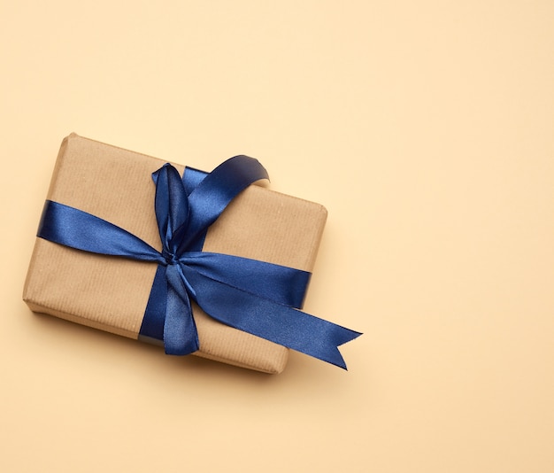 Box wrapped in brown paper and tied with a blue silk ribbon with a bow, gift on beige, top view