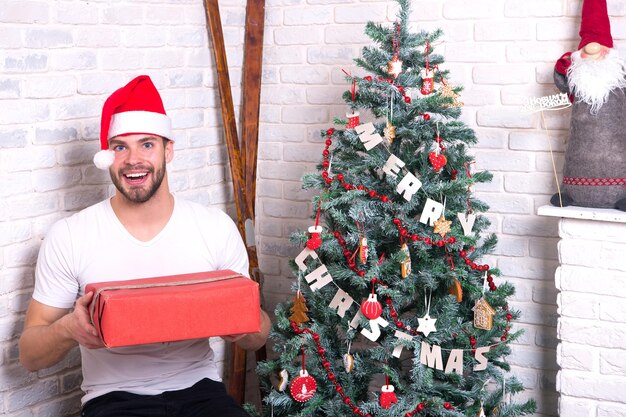 Boxing day concept. Macho in red hat hold wrapped presents. Gift giving and exchange. Man santa smile with boxes at christmas tree. Holidays preparation and celebration.