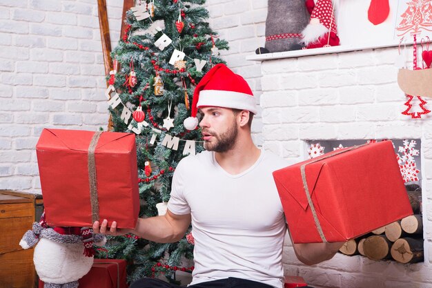 Boxing day concept. Man with red boxes at xmas tree. Merry christmas and happy new year. Macho in santa hat hold wrapped gifts at fireplace. Holidays preparation and celebration.