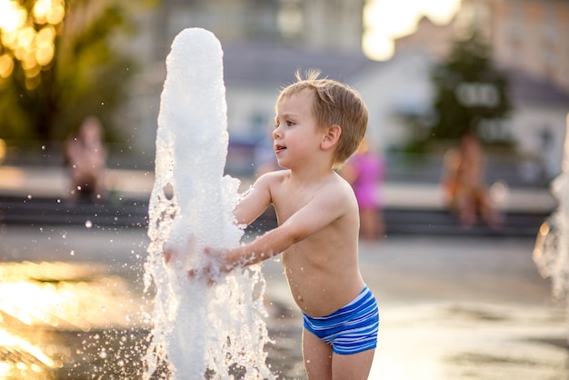 a boy of 4 years old dressed in swimming trunks and crocs plays with splashes of fountain