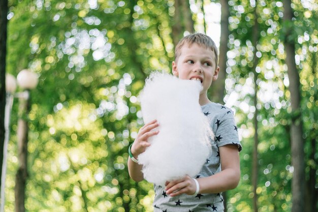 Boy 7-10 eating cotton candy in sunny park, among tall trees on green grass