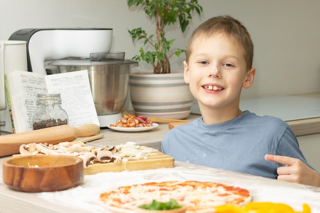 Boy 7-10 in t-shirt cooking pizza in kitchen, kid smiles and points finger at pizza