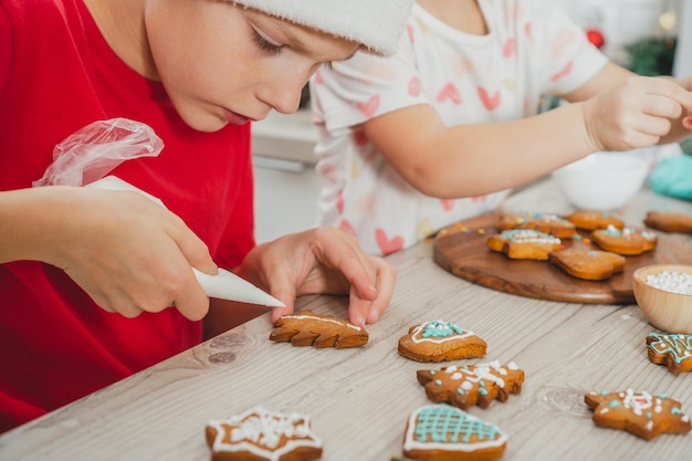 Boy 8 years old in red Santa hat decorate Christmas gingerbread cookies with icing on wooden table in kitchen