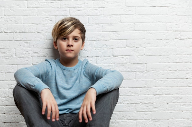 A boy of 9-10 years old sits against a background of a white brick wall. Space for text.