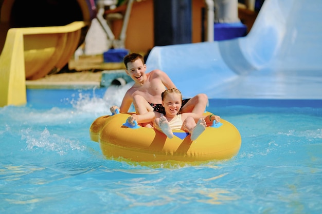 Фото Мальчик и девочка весело катаются на водной горке в аквапарке дети плавают на желтом надувном плоту.