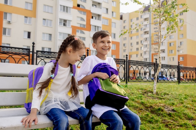 Фото Школьники мальчика и девочки сидят на скамейке с яркими рюкзаками и улыбаются, смотрят в камеру