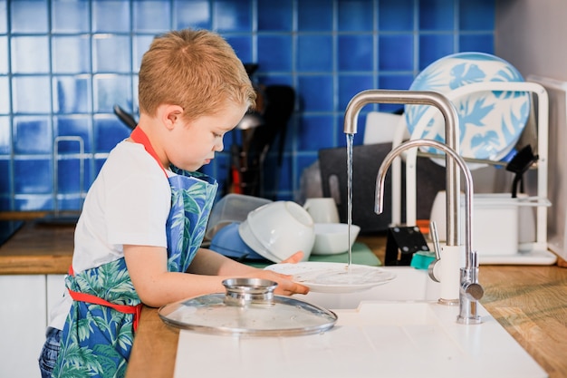 A boy in an apron washes dishes in the kitchen at home