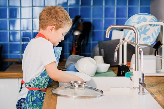 A boy in an apron washes dishes in the kitchen at home.