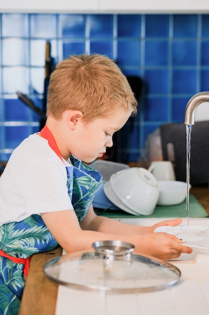 Photo a boy in an apron washes dishes in the kitchen at home.