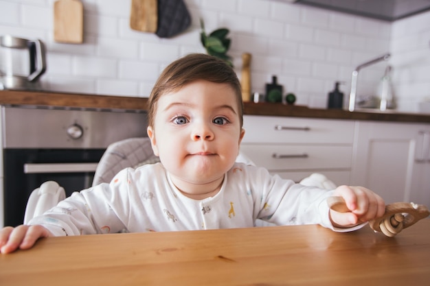Boy baby sitting in high chair and laughing in a modern white kitchen. Healthy nutrition for kids. Cute toddler side view