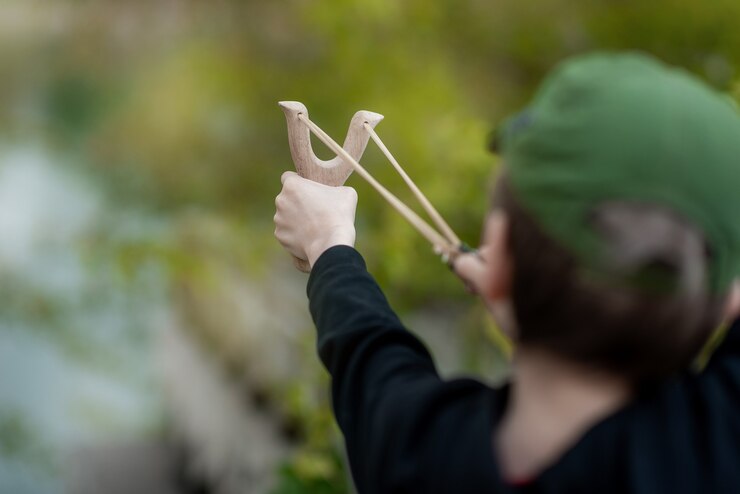 A boy in a black jacket with a hood and a cap shoots from a slingshot blurred background
