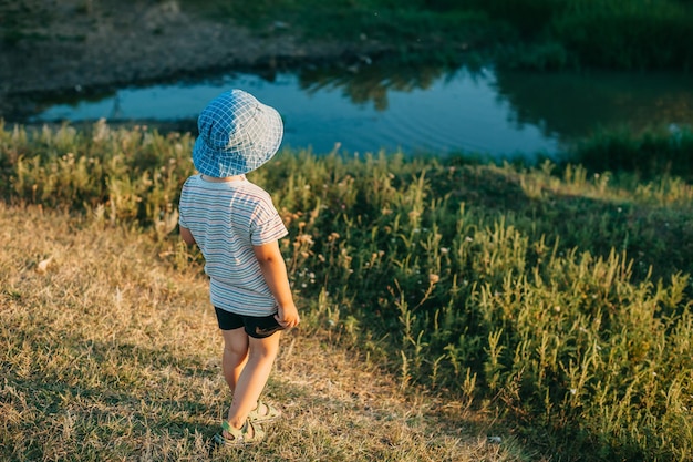 A boy in a blue hat sitting on the shore of a reservoir looking at lake in park