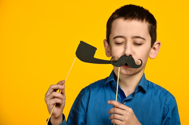 A boy in a blue shirt on a yellow background grimaces and dresses a mustache and holds a pipe