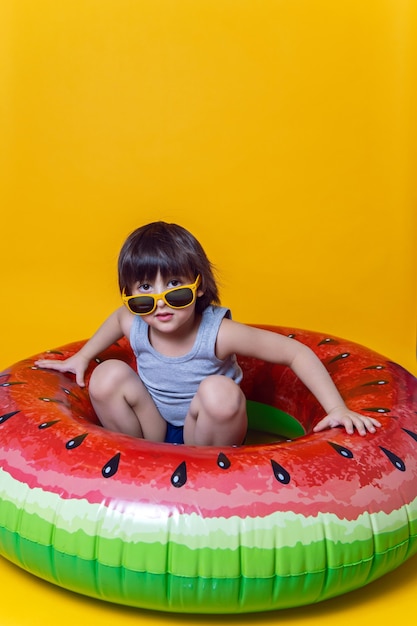 Boy in blue swimming trunks and sunglasses lying on an inflatable circle in the form of a watermelon on a yellow wall