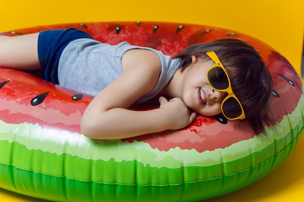 Boy in blue swimming trunks and sunglasses lying on an inflatable circle in the form of a watermelon on a yellow wall