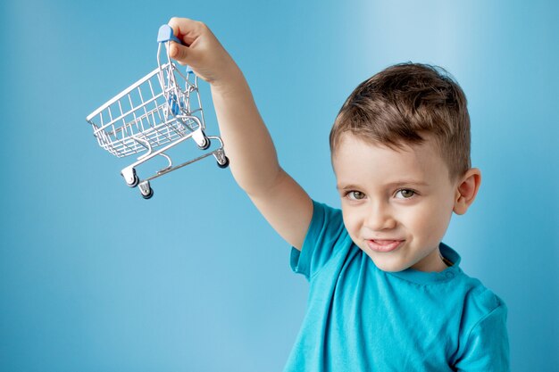 Boy in blue t-shirt is holding little cart for buying goods, products on blue