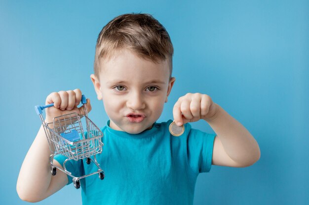 Boy in blue t-shirt is holding shopping cart and coin on blue background. Shopping and sale concept.