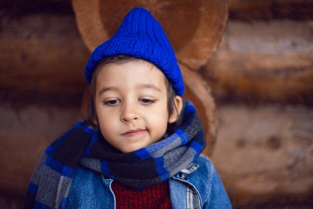 Boy child in a blue denim jacket and hat is sitting at a wooden house in autumn