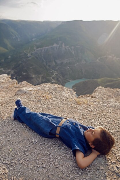 Boy child in a blue linen jumpsuit lies on the cliff of the Sulak canyon in Dagestan and thinks about life