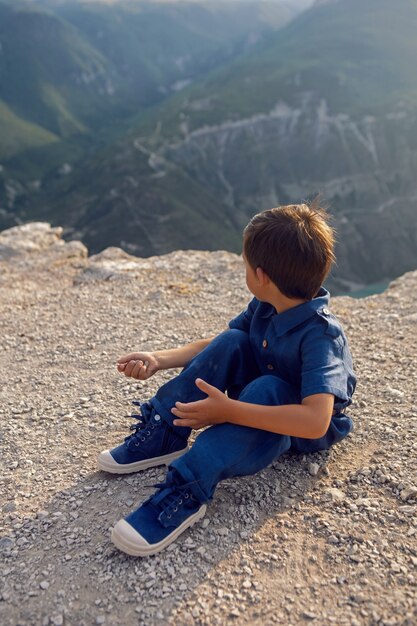 Boy child in a blue linen jumpsuit sit on the cliff of the Sulak canyon in Dagestan and thinks about life