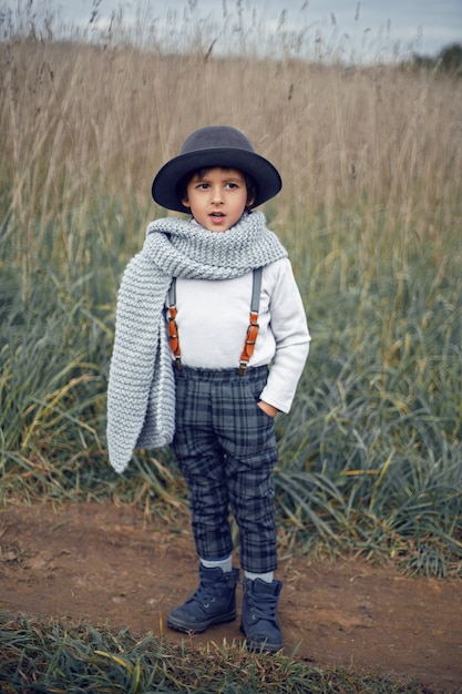 Boy child in plaid pants, hat, suspenders and scarf stands in a field