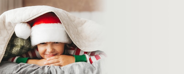 Boy child in a Santa hat and pajamas lies on a bed with white linens new year christmas