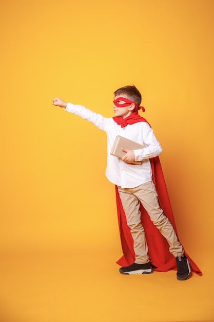 Boy child schoolboy with glasses hol book in his hands in a super hero costume on yellow background