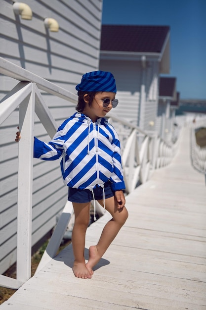 Boy child in a striped blue sailor jacket walks on the street among white wooden houses by the sea