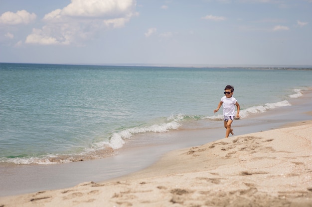 Un bambino in pantaloncini a righe e una maglietta bianca cammina su una spiaggia sabbiosa e indossa occhiali da sole