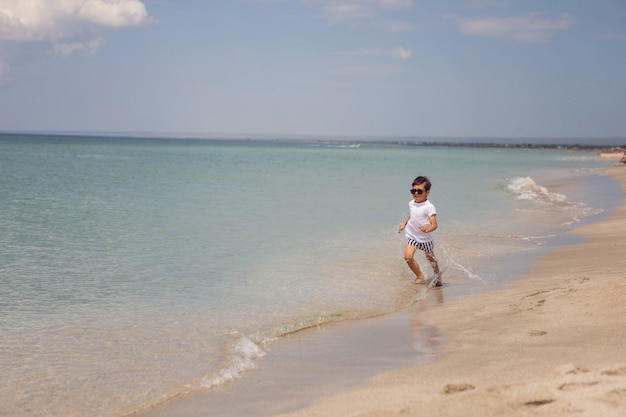 Boy child in striped shorts and a white T-shirt walks on a sandy beach and in sunglasses