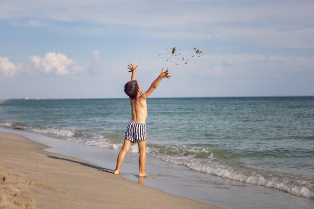 Мальчик в полосатых плавках бросает песок на пляж у моря