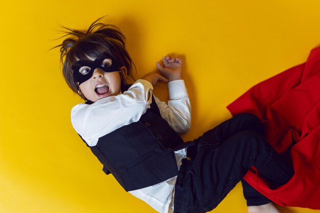 Photo boy child super hero in a suit and black mask in the studio on a yellow background barefoot