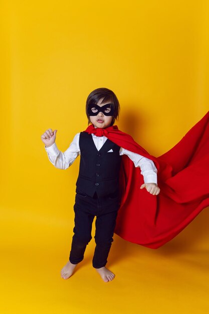 Photo boy child super hero in a suit and black mask in the studio on a yellow background barefoot