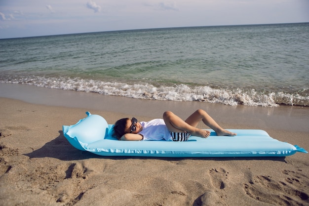 Boy child in a white T-shirt and striped swimming trunks and sunglasses is lying on a blue inflatable mattress on the beach the sailor